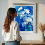 A woman in a white sweater stands looking at a large, white-framed painting of white flowers in a blue jar against a textured blue background, hanging above a light wood console table in a bright room.