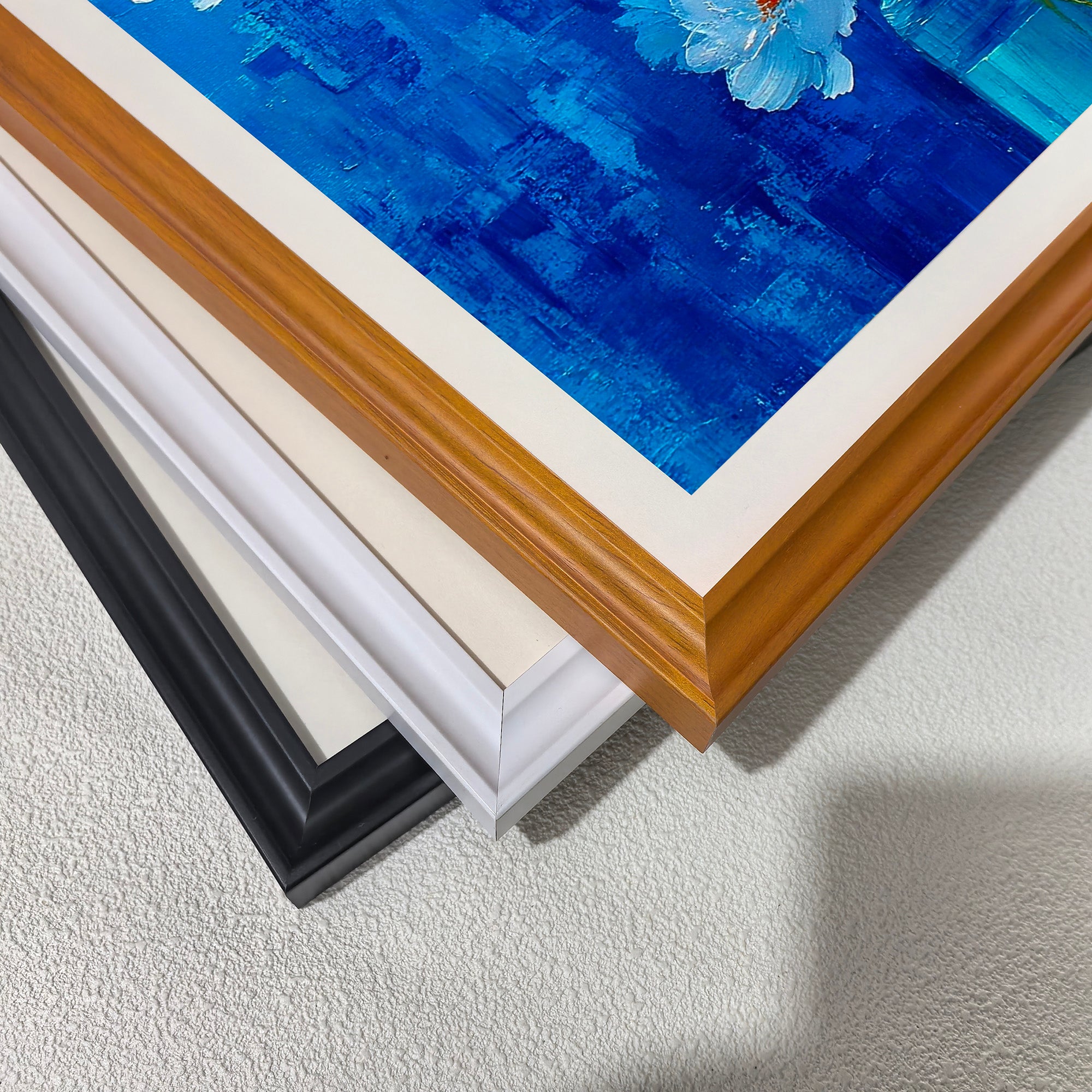A close-up, stacked view of the corners of three different wooden print frames: black, white, off-white, and light brown (wood grain), placed over a section of the painting's blue background.