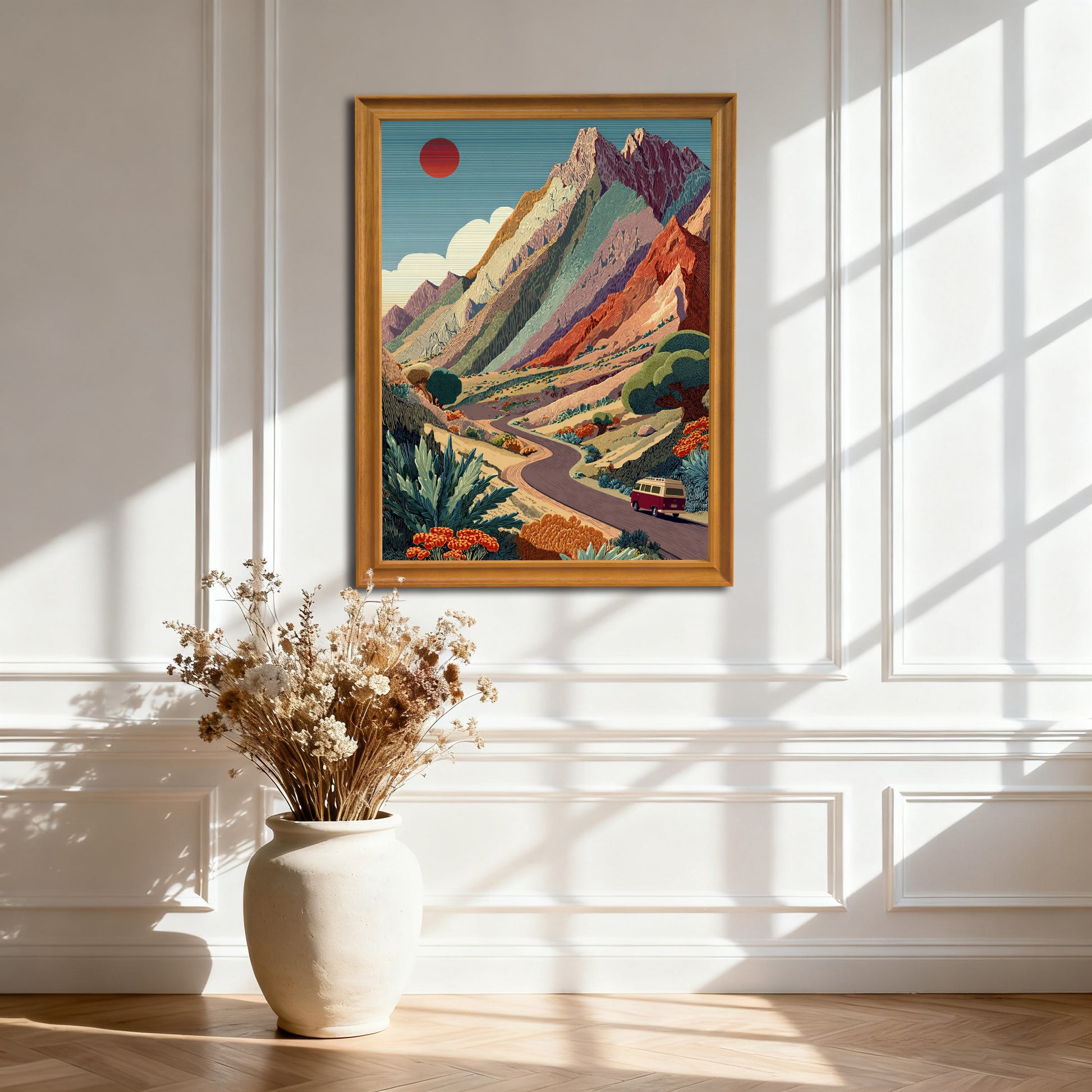 Natural wood-framed art print of the colorful mountain road trip scene, hanging on a white paneled wall. Sunlight casts dramatic geometric shadows.