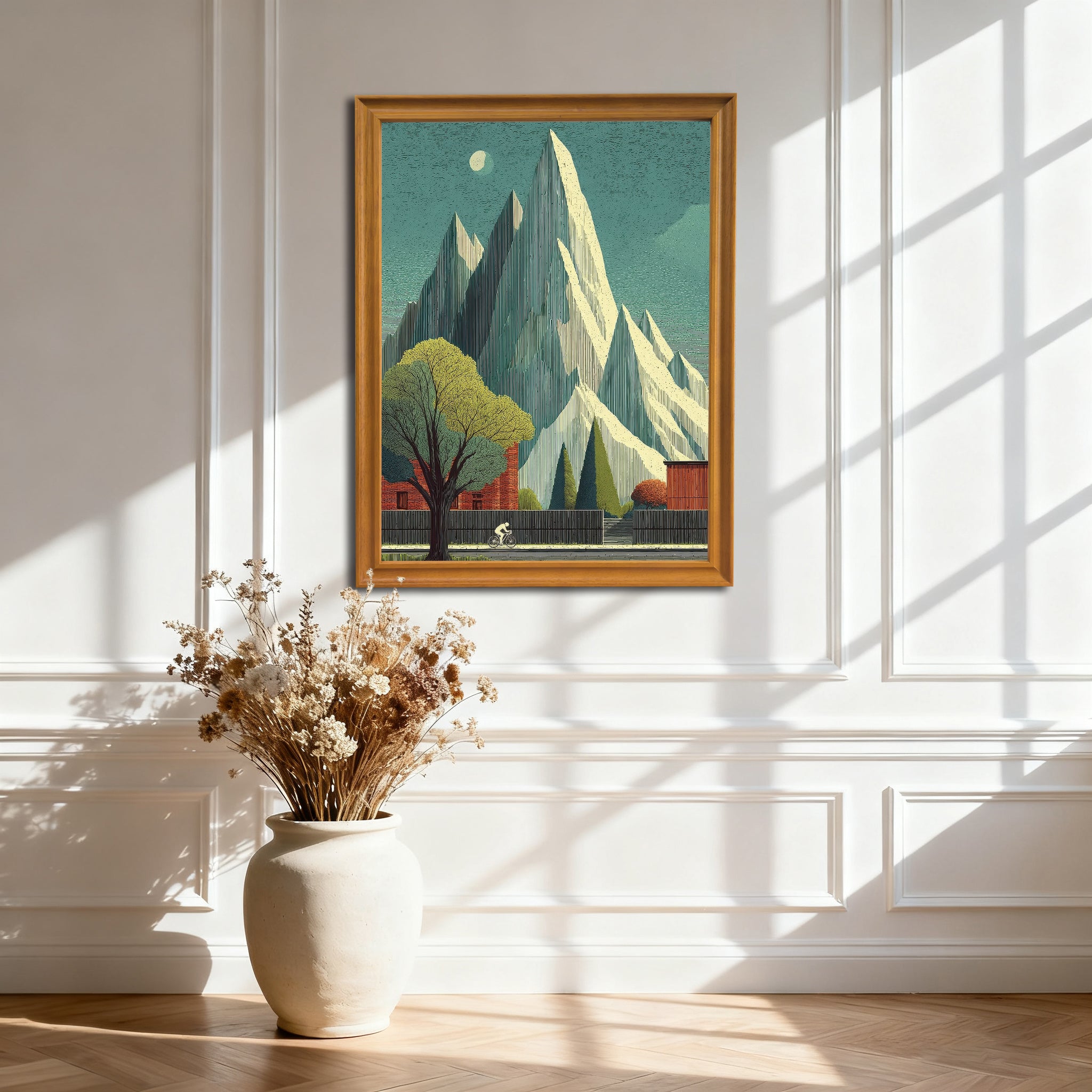 Natural wood-framed art print featuring sharp white mountains and a cyclist passing a large tree. The print is hanging on a white paneled wall with dramatic sunlight and shadows.