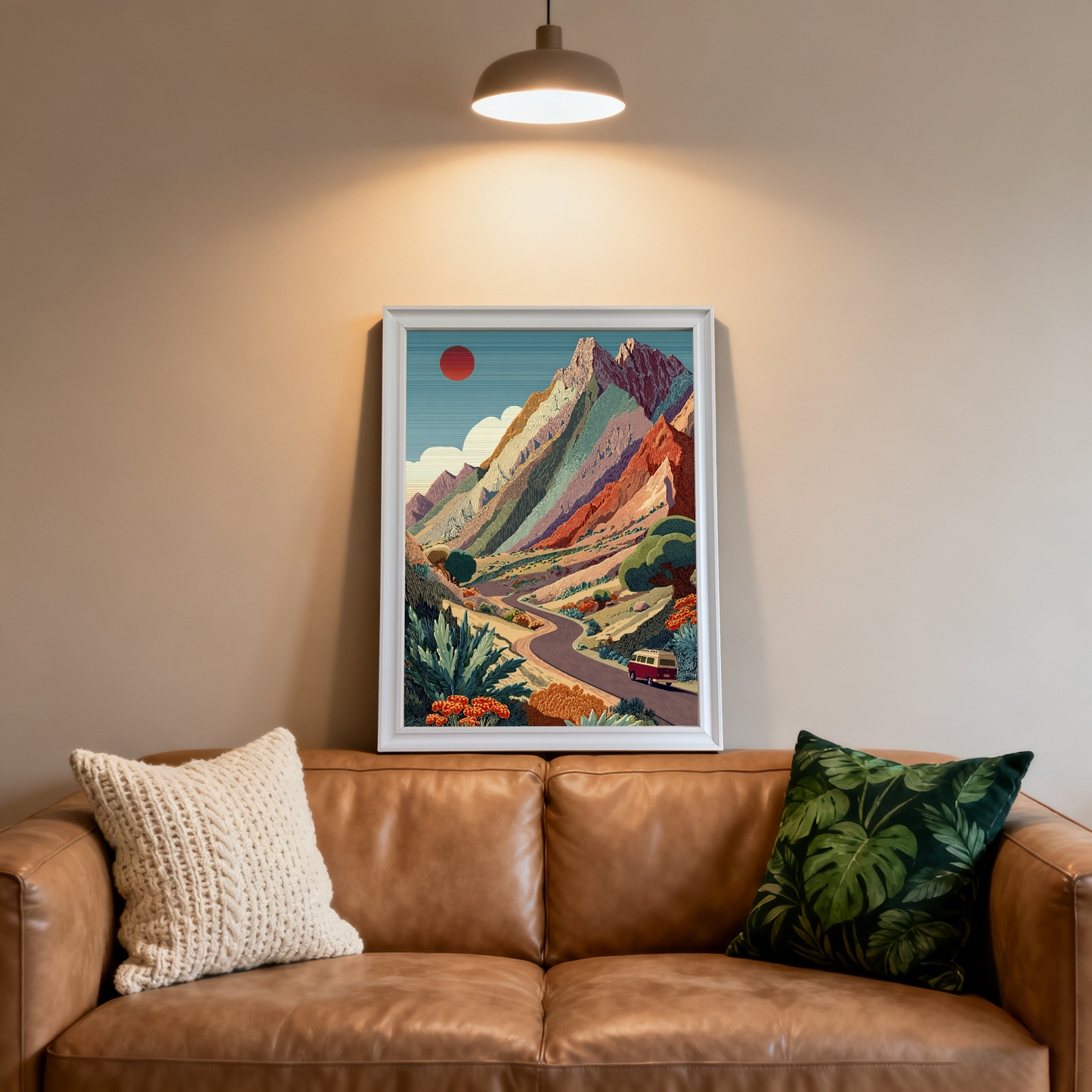 White-framed art print featuring the colorful mountain road trip scene, centered above a brown leather couch and highlighted by a ceiling light.