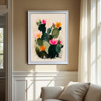 The abstract blooming cactus art print, framed in a contemporary white frame, is hung in a living room with beige walls and large windows, showcasing the artwork's bright colors over a white sofa.