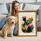 A smiling woman sits on the floor next to a golden retriever puppy, holding a framed art print. The artwork is an abstract illustration of a dark green, blooming cactus with vibrant pink and orange flowers, set in a wood grain frame.
