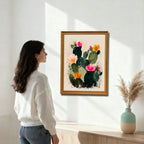 A woman in a white sweater looking at a framed art print of an abstract prickly pear cactus with pink and orange flowers, set on a cream background, hanging on a white wall in a gold/natural wood frame.