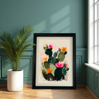 The abstract blooming cactus art print, framed in a thick black frame, is leaning against a dark teal paneled wall next to a large potted palm plant and a bright window.