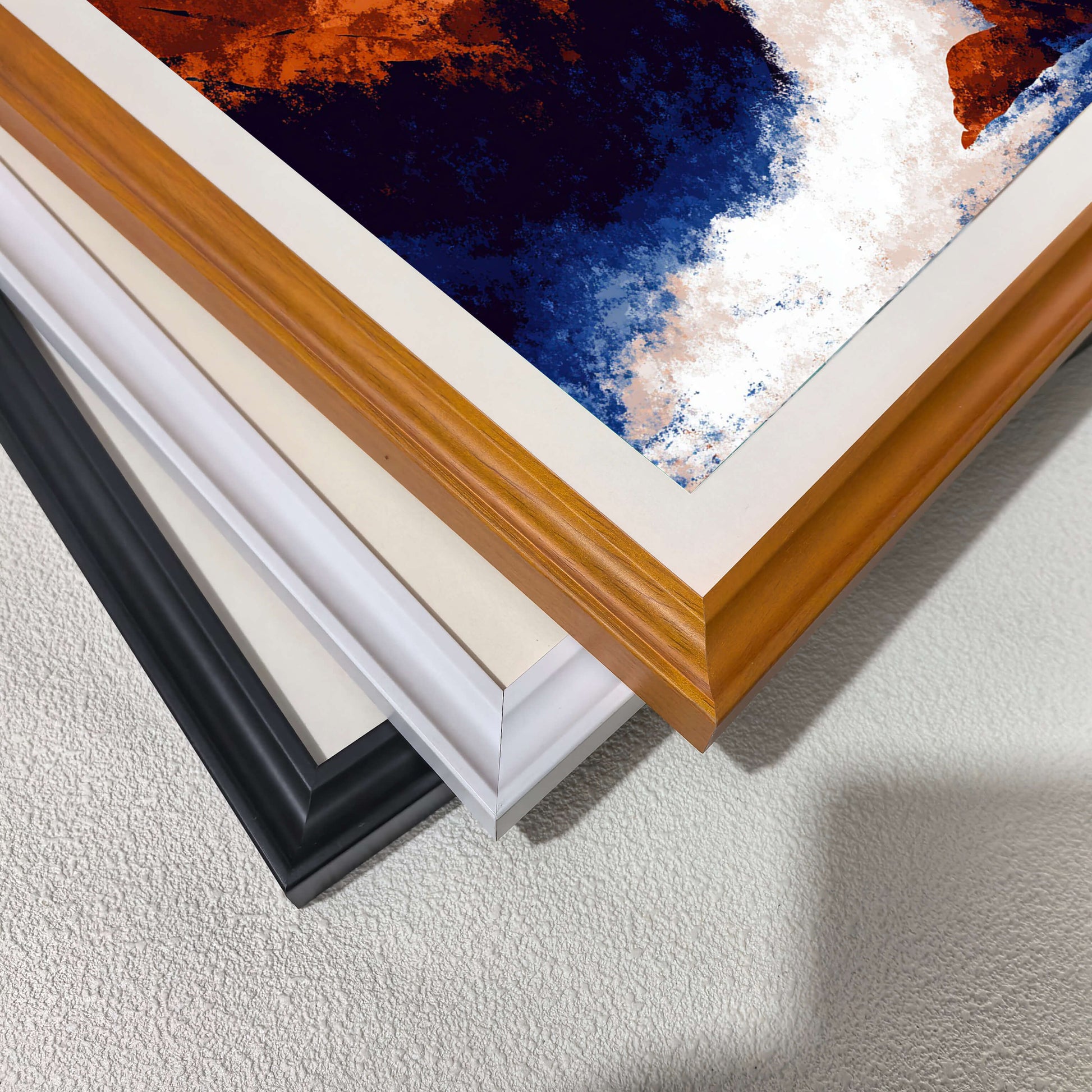 Stacked close-up view of the three different wood frame options: black, white,  and dark wood (brown/natural), with a corner of the waterfall art print visible.