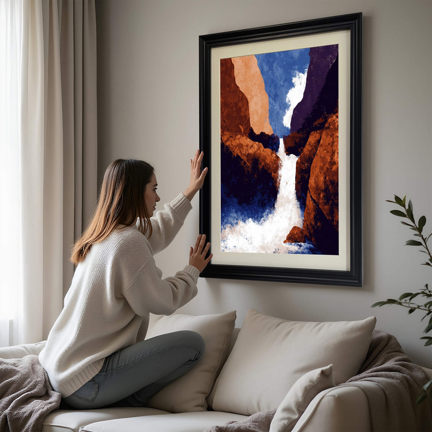 A woman in a white sweater adjusting a large abstract art print of a waterfall in a canyon, framed in black, hanging above a beige sofa.