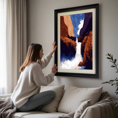 A woman in a white sweater adjusting a large abstract art print of a waterfall in a canyon, framed in black, hanging above a beige sofa.