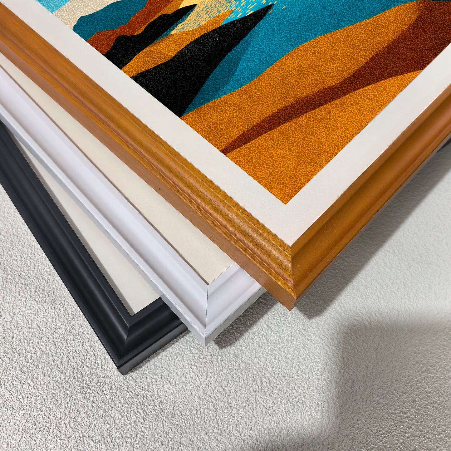 Close-up stack of three wood picture frames—black, white, and natural light brown wood—displaying a stippled abstract landscape print with blue water, orange/yellow land, and black rocks, highlighting framing choices.