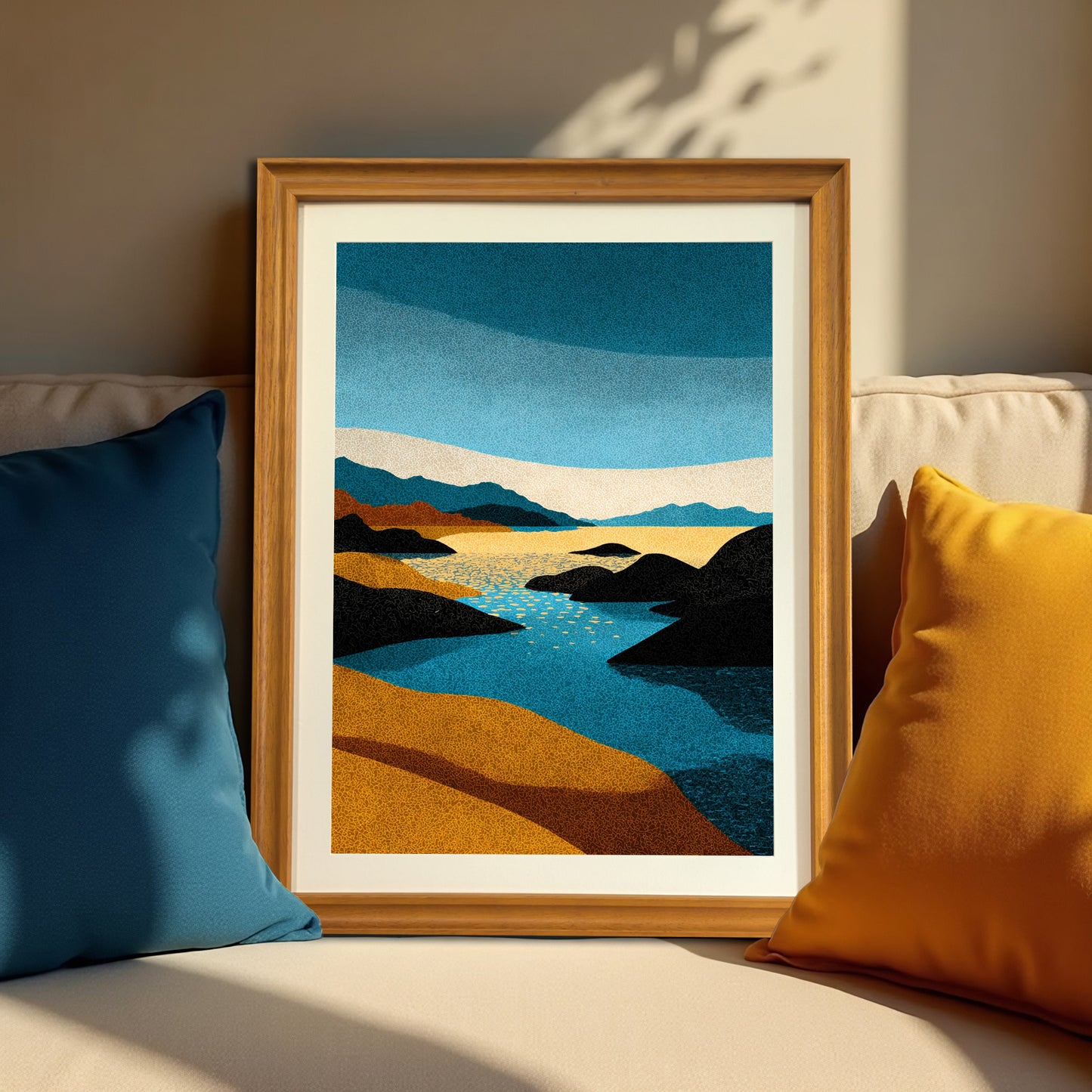Vertical abstract stippled landscape art print in a natural brown wood frame, resting on a neutral-colored couch between a dark blue and a bright yellow throw pillow, highlighted by sun and shadow.