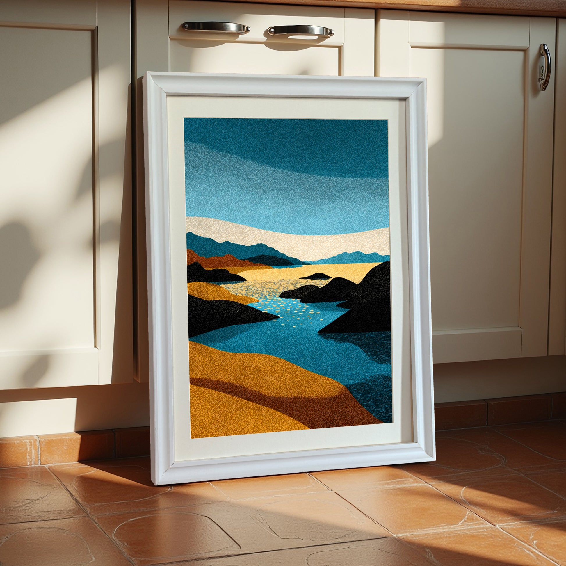 Vertical abstract landscape art print with blue, yellow, and black stippled texture, framed in a thick white wooden frame with a cream mat, leaning against cream kitchen cabinets on a terracotta tile floor.