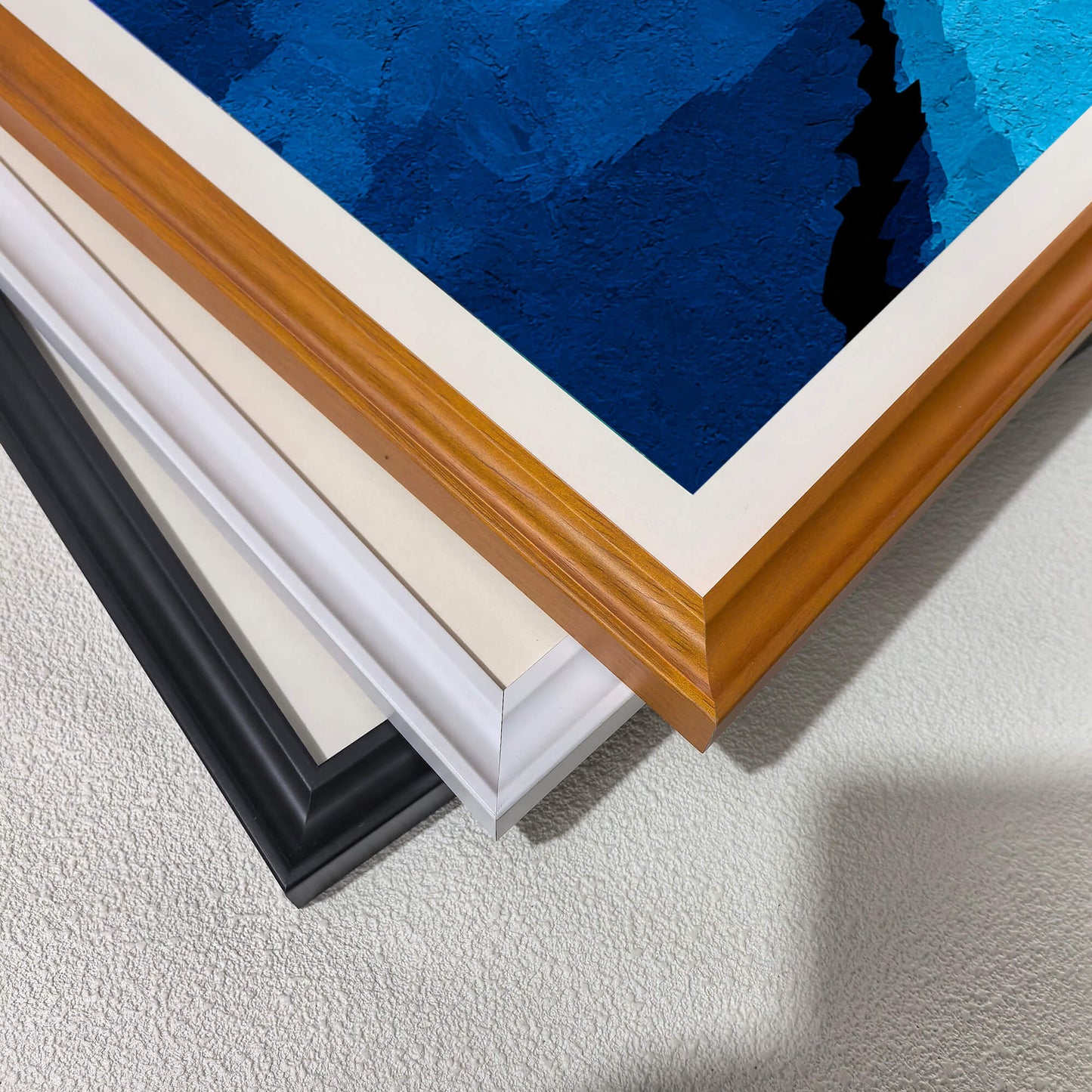 Stacked close-up view of the three different wood frame options: black, white, and dark wood (brown/natural), highlighting the texture of the blue print.