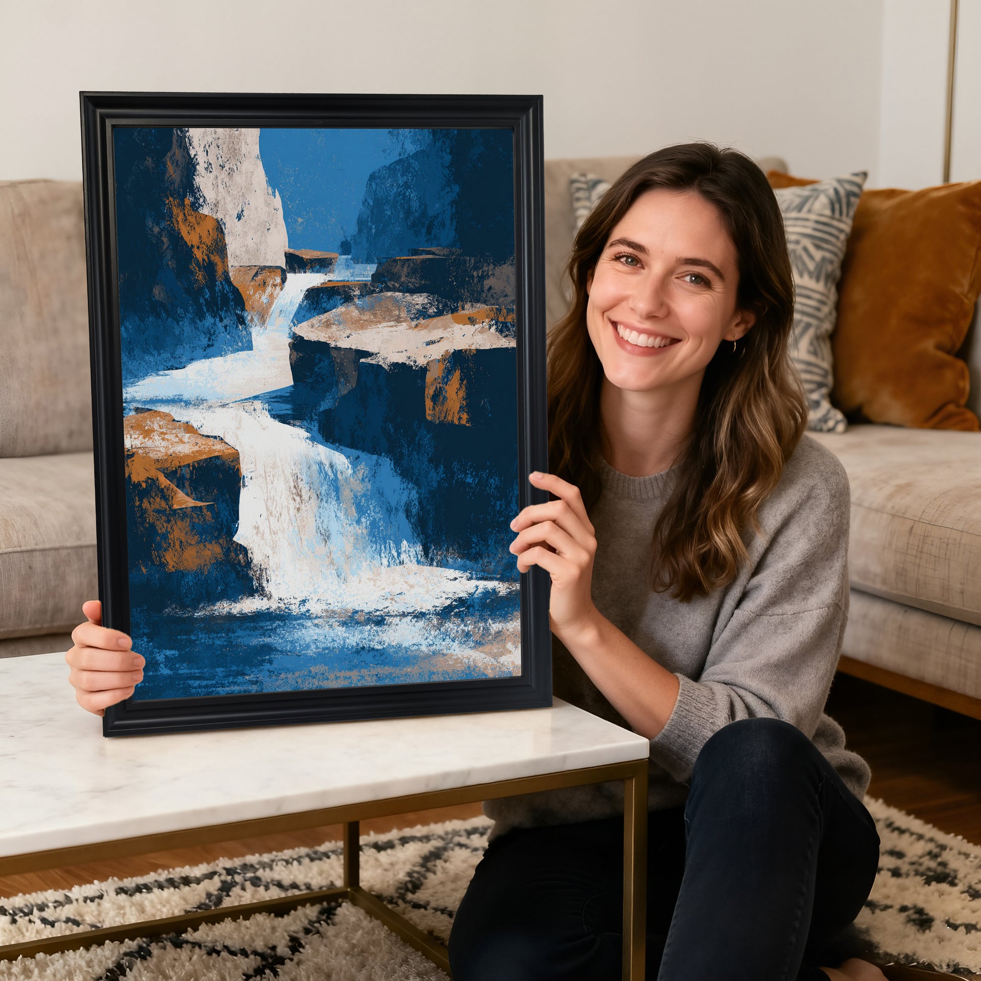 A smiling woman sitting on the floor holding a vertical abstract waterfall art print in a thick black wood frame, with a white coffee table and sofa in the background.