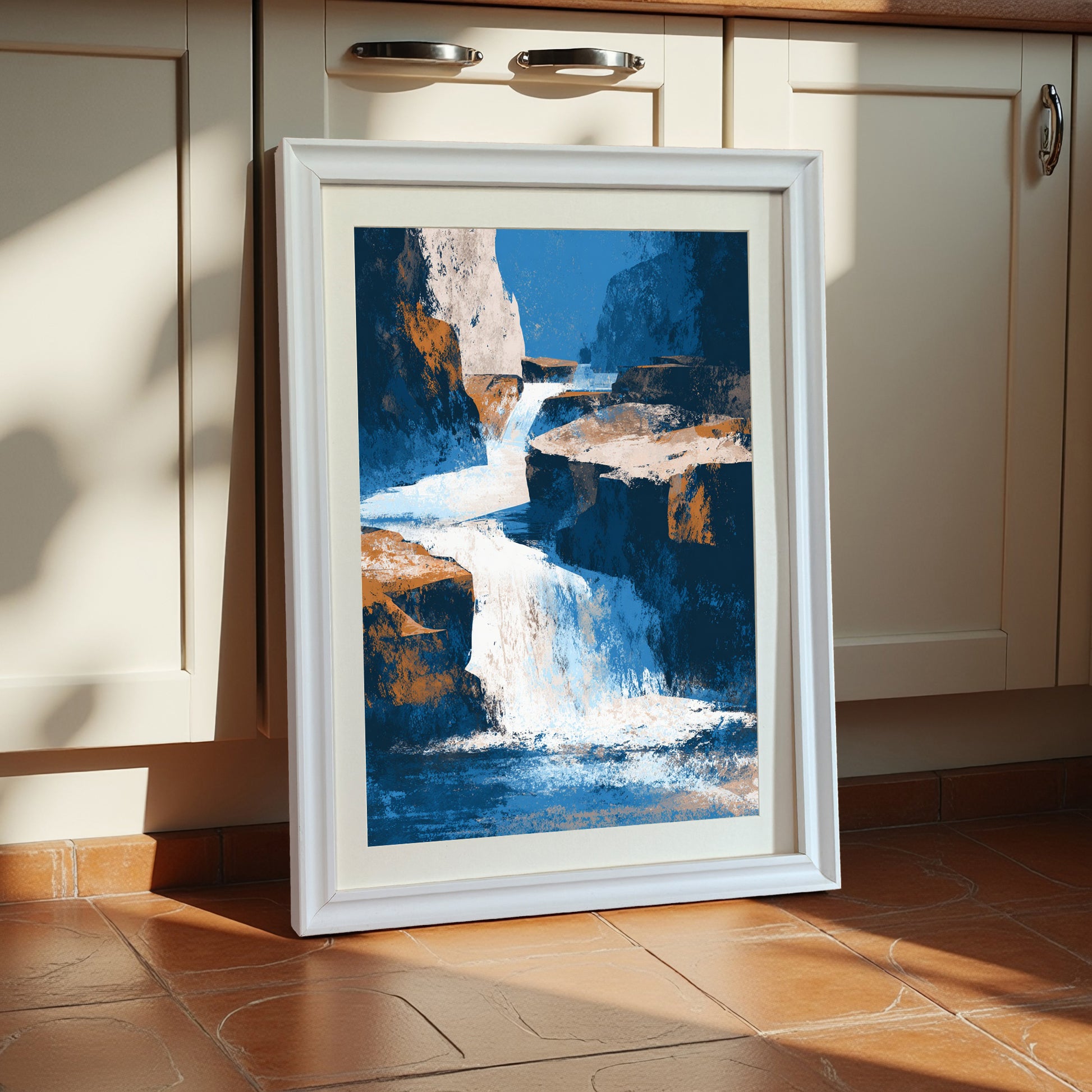 Vertical abstract waterfall art print with blue, white, and orange colors, framed in a thick white wooden frame with a cream mat, leaning against cream kitchen cabinets on a terracotta tile floor.