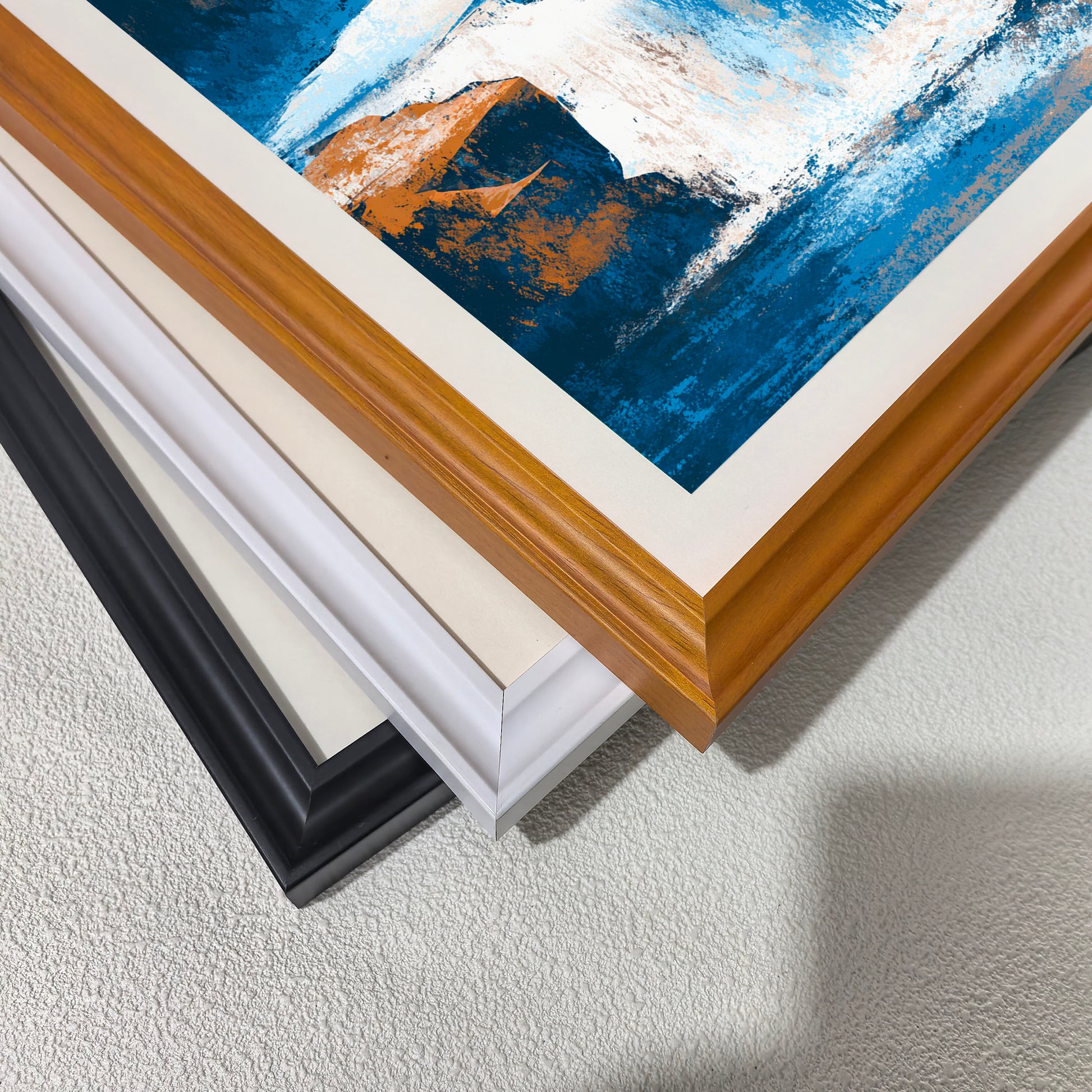 Close-up stack of three wood picture frames—black, white, and natural light brown wood—displaying a painting with abstract blue, white, and orange brushstrokes, suggesting different framing options.