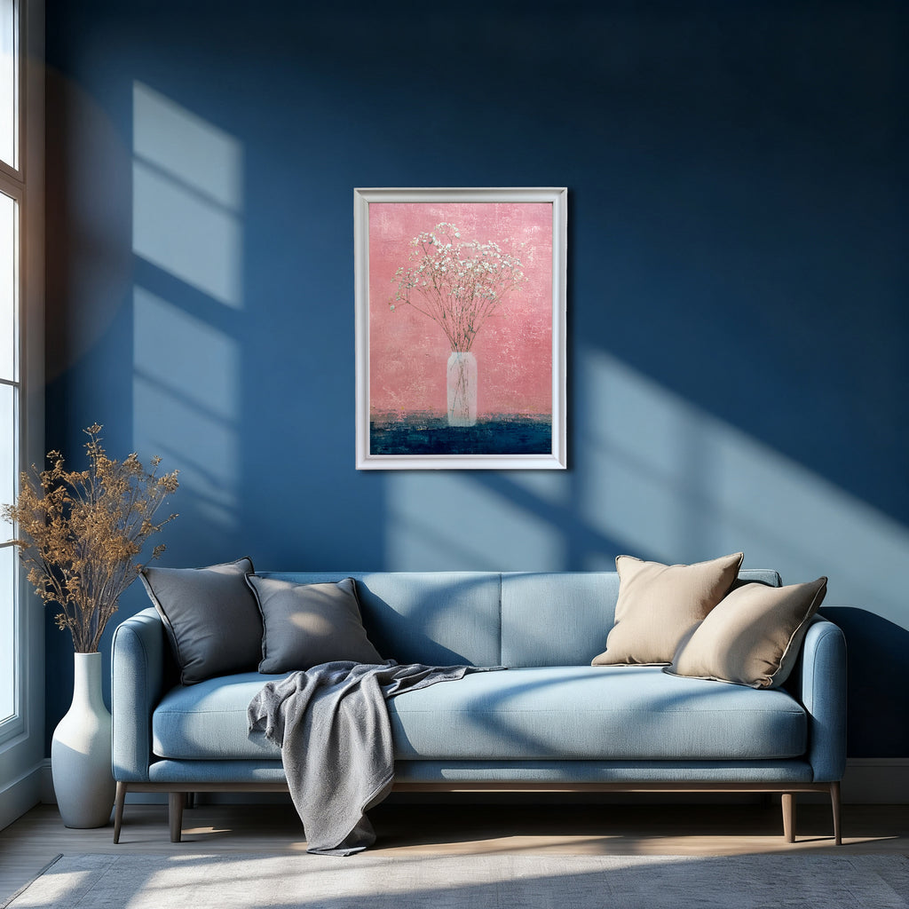 White-framed art print of baby's breath in a vase against a pink and dark blue background, hanging above a light blue sofa on a deep navy blue wall with bright window light.