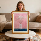 Smiling woman holding up a framed art print of baby's breath in a vase with a gold-colored wood frame, sitting behind a round marble coffee table.