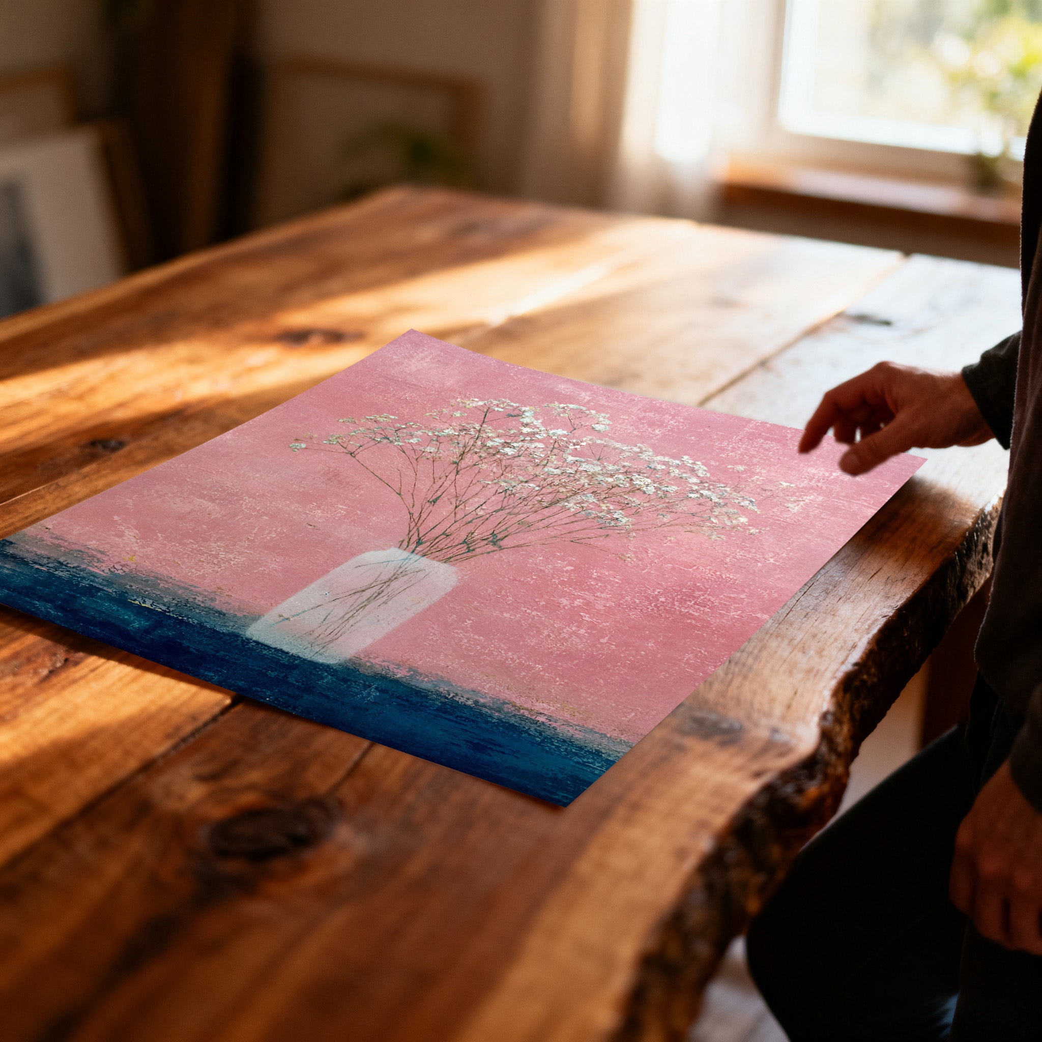 Unframed art print of baby's breath in a vase on a textured pink and dark blue background, lying on a rustic wooden table with a person's hand reaching toward it.