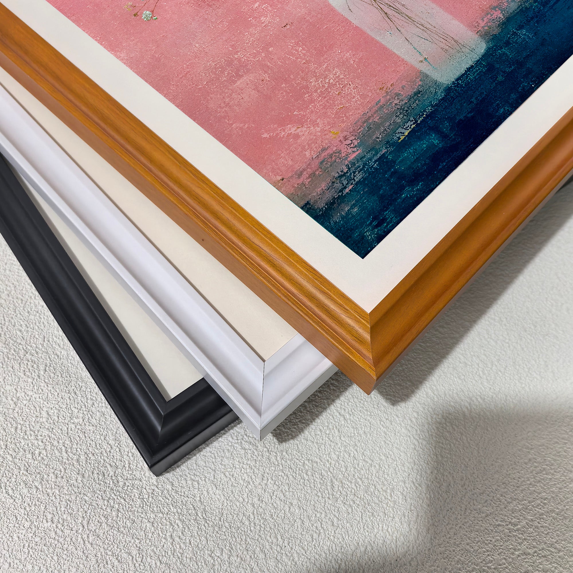 Close-up view of three stacked wood picture frame corners in black, white, and natural brown, showing the art print inside which features vibrant pink and dark blue colors.