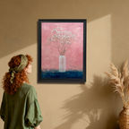 Woman with red hair looking at a black-framed art print of baby's breath in a vase, hanging on a warm beige wall with a wicker vase nearby.