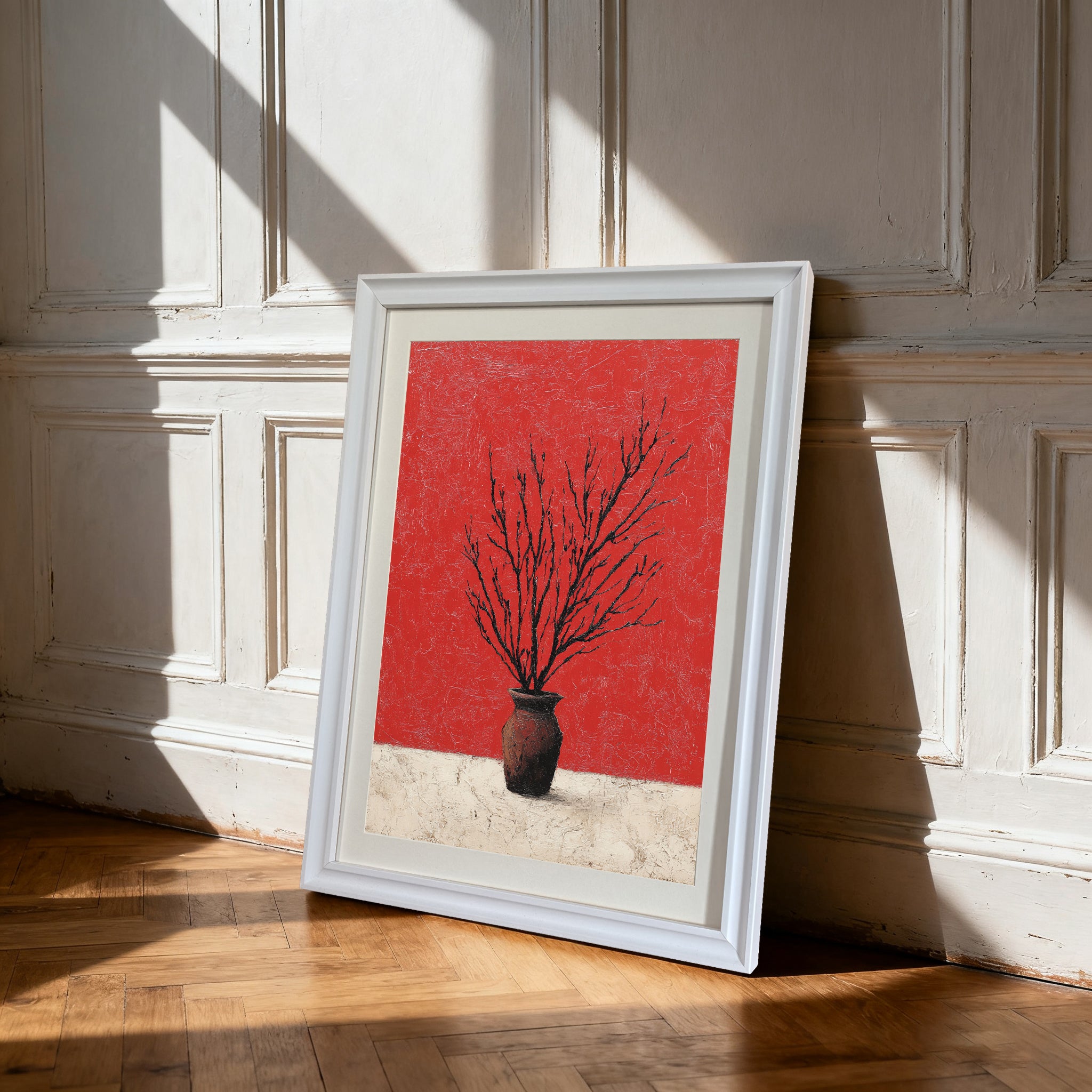 White-framed art print of branches in a vase against a red and textured white background, leaning on a hardwood floor against a white paneled wall with strong sun shadows.