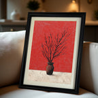 Black-framed art print of dark branches in a vase against a red and textured white background, leaning on a light-colored sofa cushion.