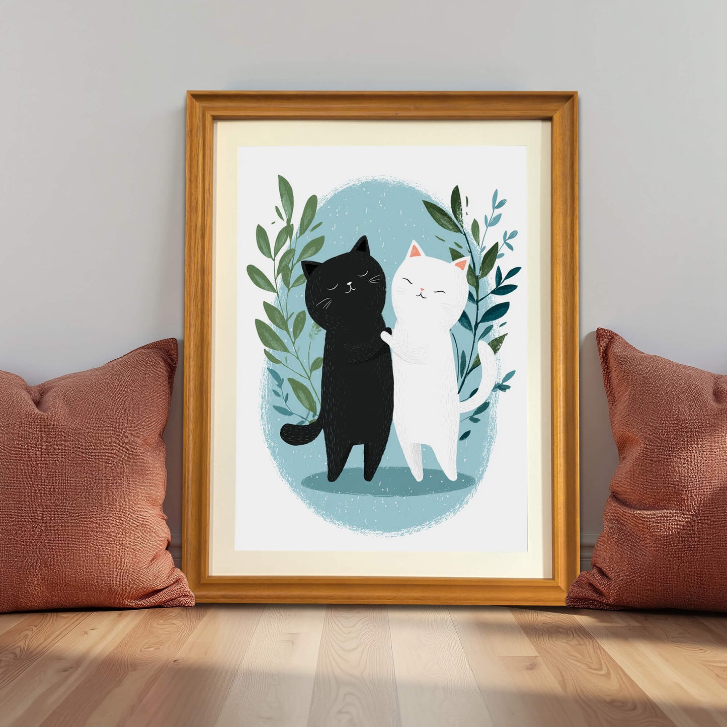 A natural wood-framed print of the cats sits on a light wood floor, leaning against a grey wall. It is flanked by two burnt-orange throw pillows, with soft sunlight casting shadows across the floor.