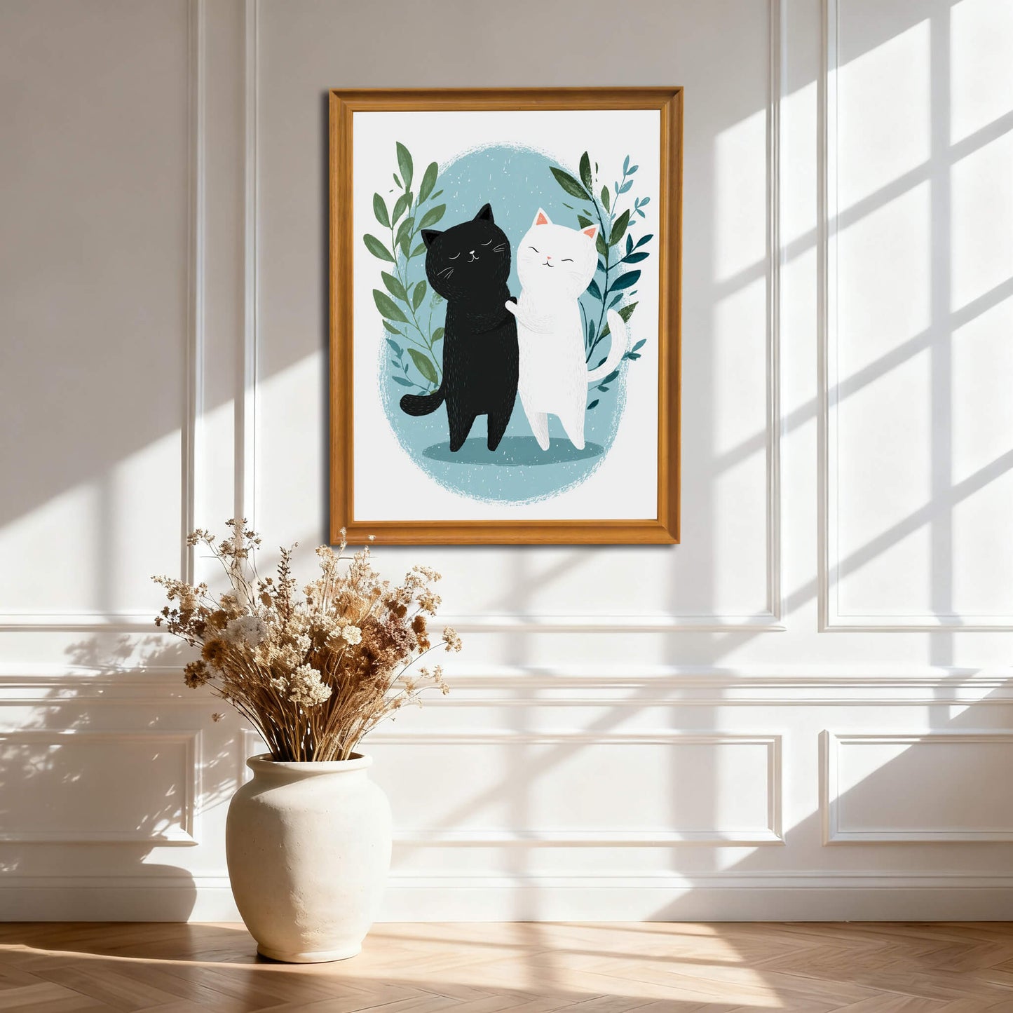A large, natural wood-framed print of the cats hangs on a white wall with decorative molding. Below it sits a large cream-colored floor vase filled with dried flowers, with sunlight creating dramatic shadows across the wall.
