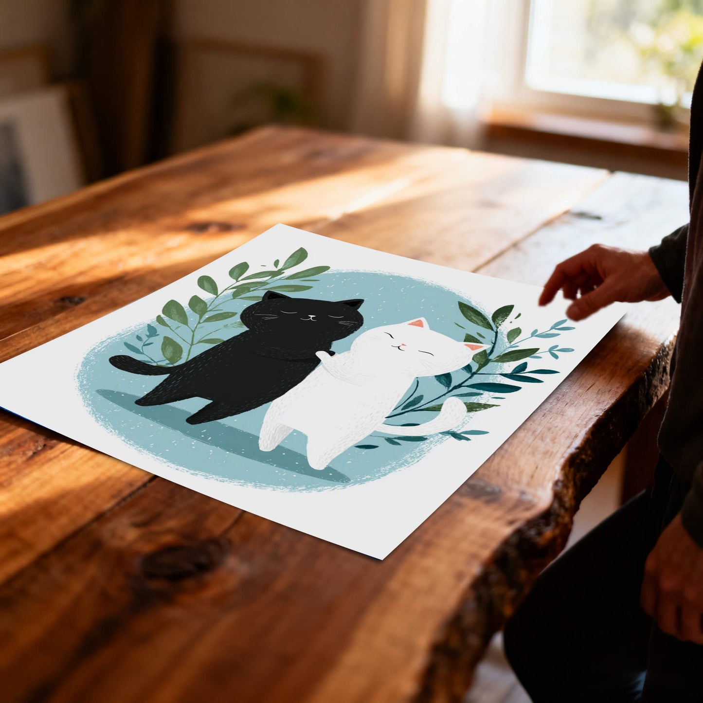 An unframed art print of the hugging cats lies flat on a rustic, live-edge wooden table. A person's hand is visible on the right side of the frame, reaching toward the paper.