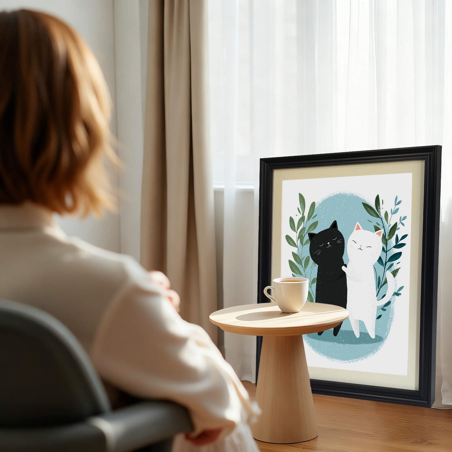 A lifestyle shot from behind a person sitting in a chair, looking toward a large black-framed print of the cats resting on the floor against a window. A small wooden side table with a cup of coffee is in the foreground.