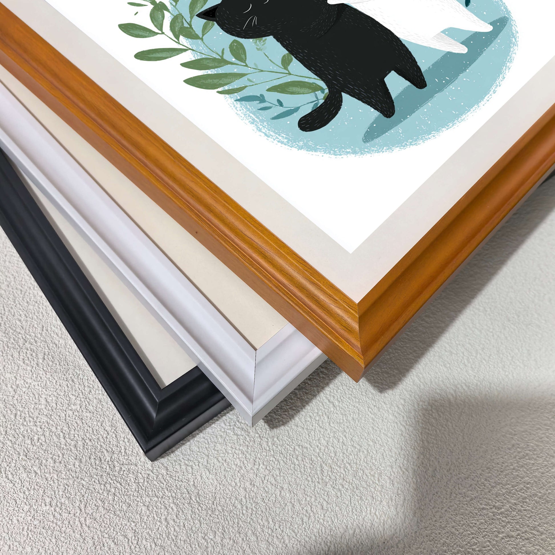 A high-angle, close-up shot of three framed art prints stacked diagonally on a textured white surface. The top frame is natural wood, followed by a white frame, and a black frame at the bottom.