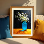A wood-framed art print resting on a beige sofa between a dark blue pillow and a yellow pillow. The artwork features a high-contrast still life of white flowers in a blue vase on an orange surface.