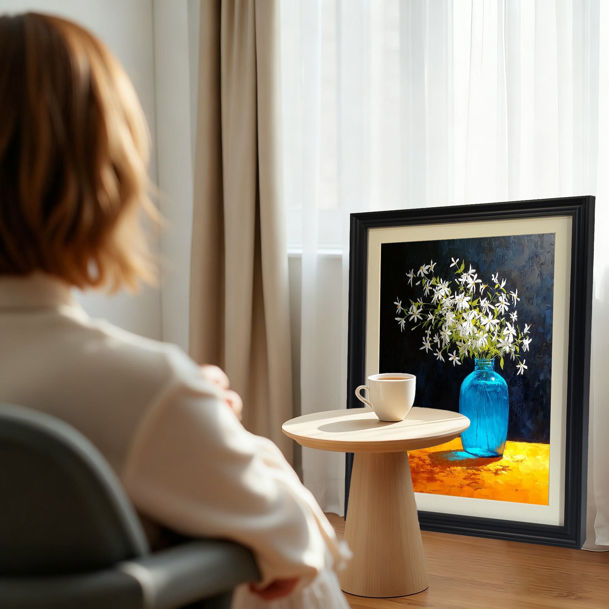 A black-framed art print leaning against a window with light curtains. The still life print is positioned next to a small wooden table with a coffee cup, with a woman looking towards it in the foreground.