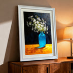 A white-framed art print leaning on a dark wood cabinet next to a glowing table lamp. The artwork is a dramatic still life of white flowers in a blue glass vase.