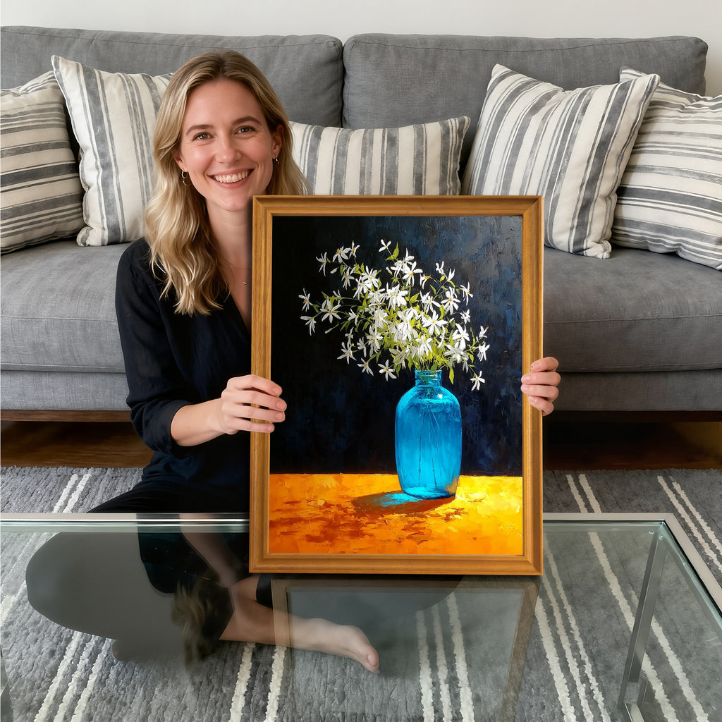 A smiling woman sitting on the floor, holding a gold-framed art print. The painting depicts white flowers in a bright blue glass vase, set against a dark background, resting on a vividly textured orange table.
