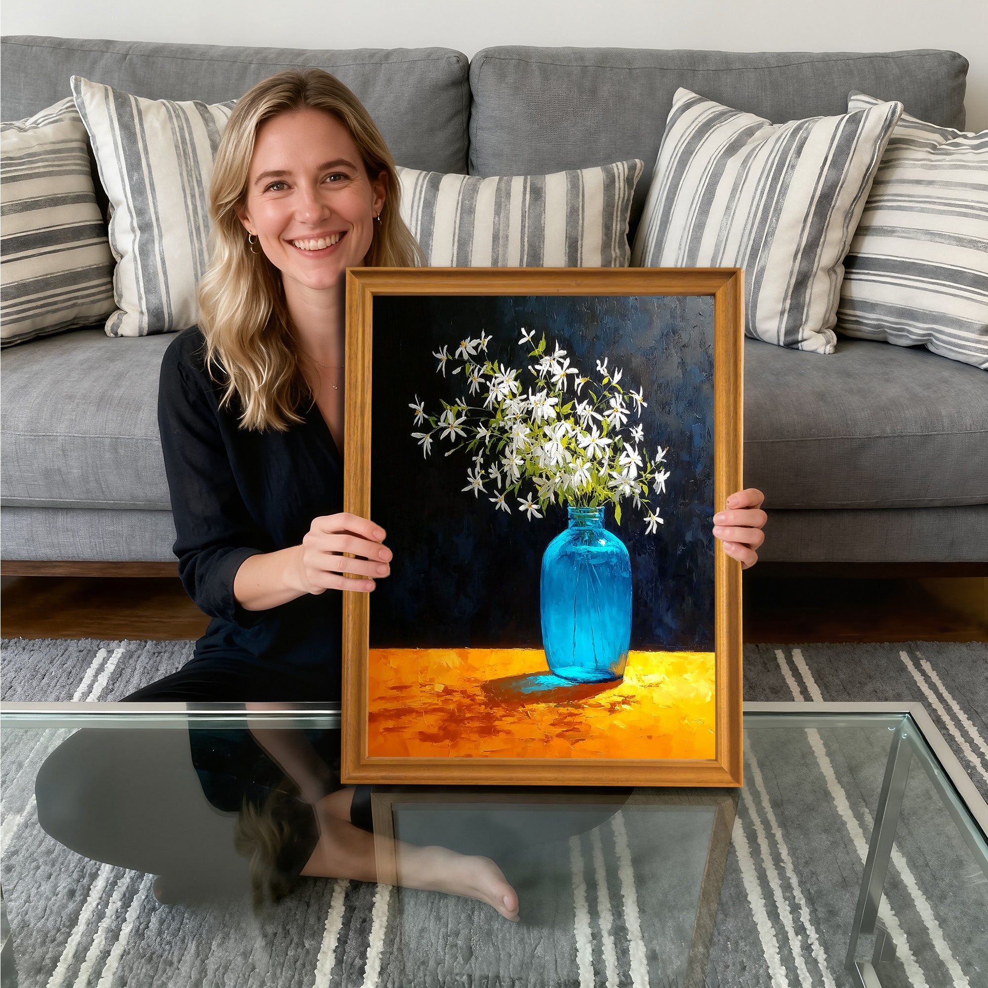A smiling woman sitting on the floor, holding a gold-framed art print. The painting depicts white flowers in a bright blue glass vase, set against a dark background, resting on a vividly textured orange table.