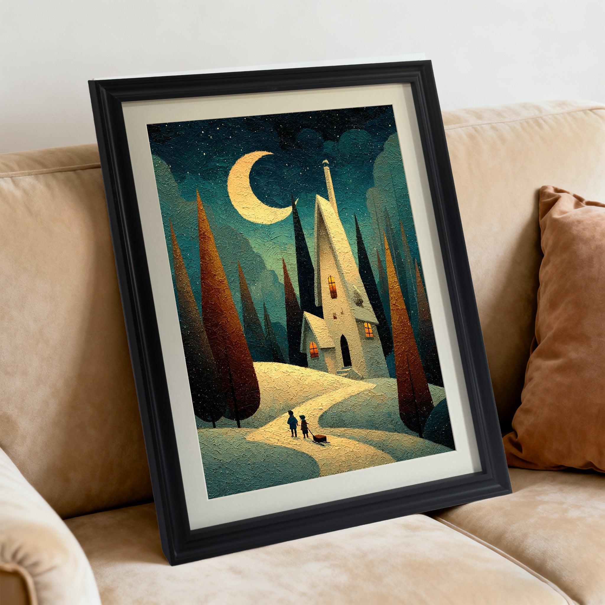Whimsical art print of a house under a crescent moon on a snowy path, framed in black, angled while resting on a tan fabric sofa with a rust-colored pillow.