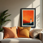 Minimalist lighthouse landscape print with an orange sky, in a black frame, resting on a white sofa between two warm orange pillows.