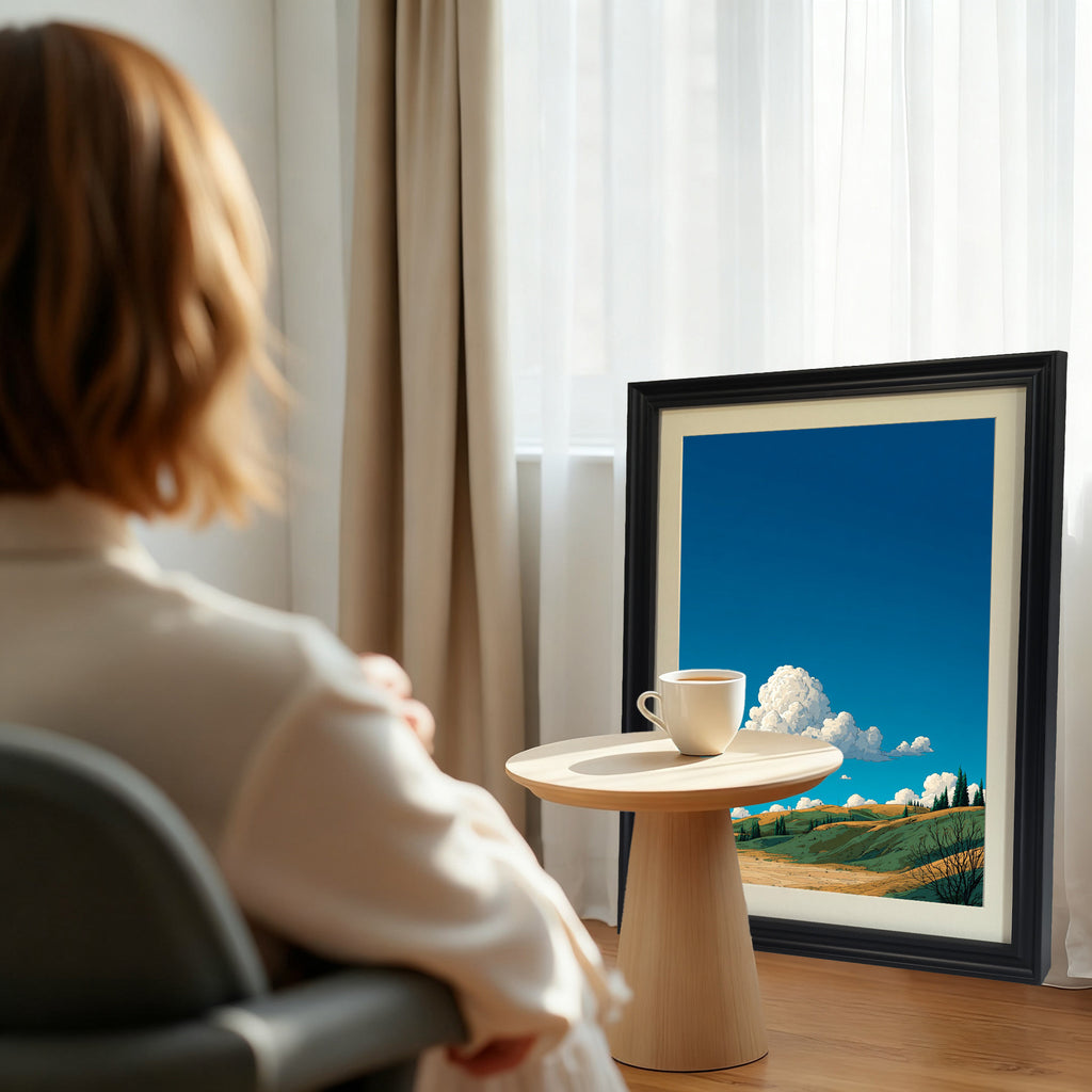 A black-framed art print leaning against a window with light-colored curtains. The artwork is a vivid landscape of hills and sky. A woman is seen from behind, looking toward the print next to a small wooden table with a coffee cup.