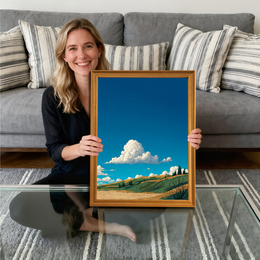 A smiling woman sitting on the floor, holding a gold-framed art print. The artwork is a vibrant, stylized landscape featuring rolling green and gold hillsides under a deep blue sky with a prominent fluffy white cumulus cloud.