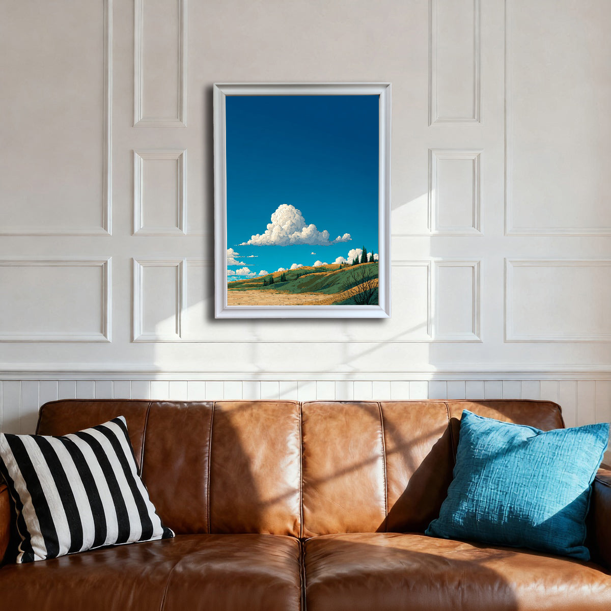 A white-framed art print centered above a brown leather sofa with striped and blue pillows. The white paneled wall provides a clean setting for the vibrant blue sky and rolling landscape artwork.