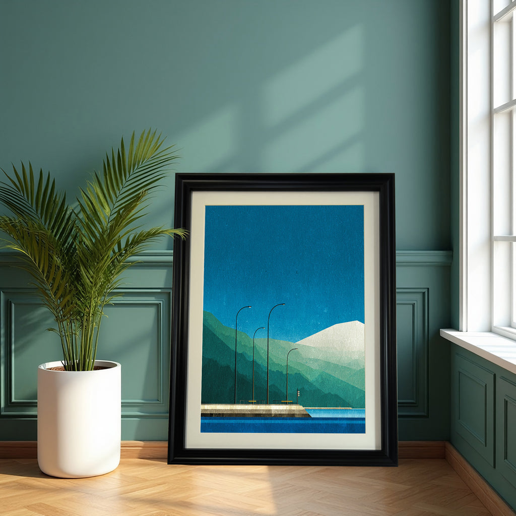 Geometric landscape art print with blue and green colors, in a chunky black frame with a mat, leaning on a wooden floor against a dark teal paneled wall next to a potted palm plant.