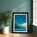 Geometric landscape art print with blue and green colors, in a chunky black frame with a mat, leaning on a wooden floor against a dark teal paneled wall next to a potted palm plant.