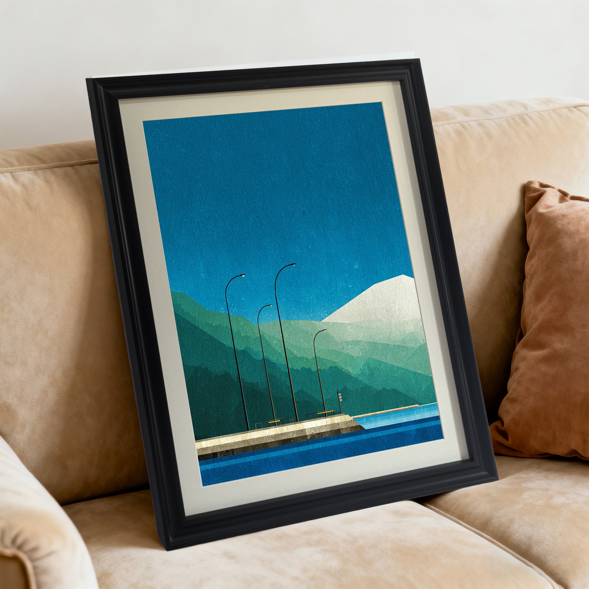 Geometric art print featuring a blue sky and layered mountains over a pier, framed in black, angled while resting on a tan fabric sofa with a rust-colored pillow.