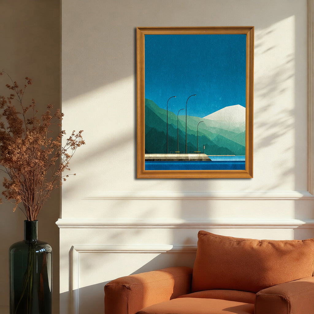 Geometric landscape art print with a blue sky, layered green mountains, and a waterfront pier with streetlights, in a gold-colored wood frame, hanging on a beige wall above an orange armchair.