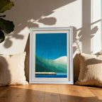Geometric art print of a blue and green mountain landscape, framed in white, leaning on a wooden floor between two cream-colored pillows by a sunlit window.