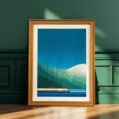 Minimalist geometric print featuring a blue sky, green mountains, and a waterfront scene, in a natural wood frame with a cream mat, leaning on a wooden floor against a dark green paneled wall.
