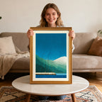 Smiling young woman holding up the geometric mountain and pier art print in a gold-colored wood frame, sitting in a living room setting with a light couch and marble coffee table.