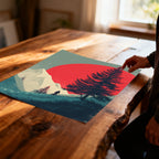 Unframed art print featuring the red sun, mountain, and biker design, lying flat on a rustic wooden table. A person's hand is reaching toward the corner.