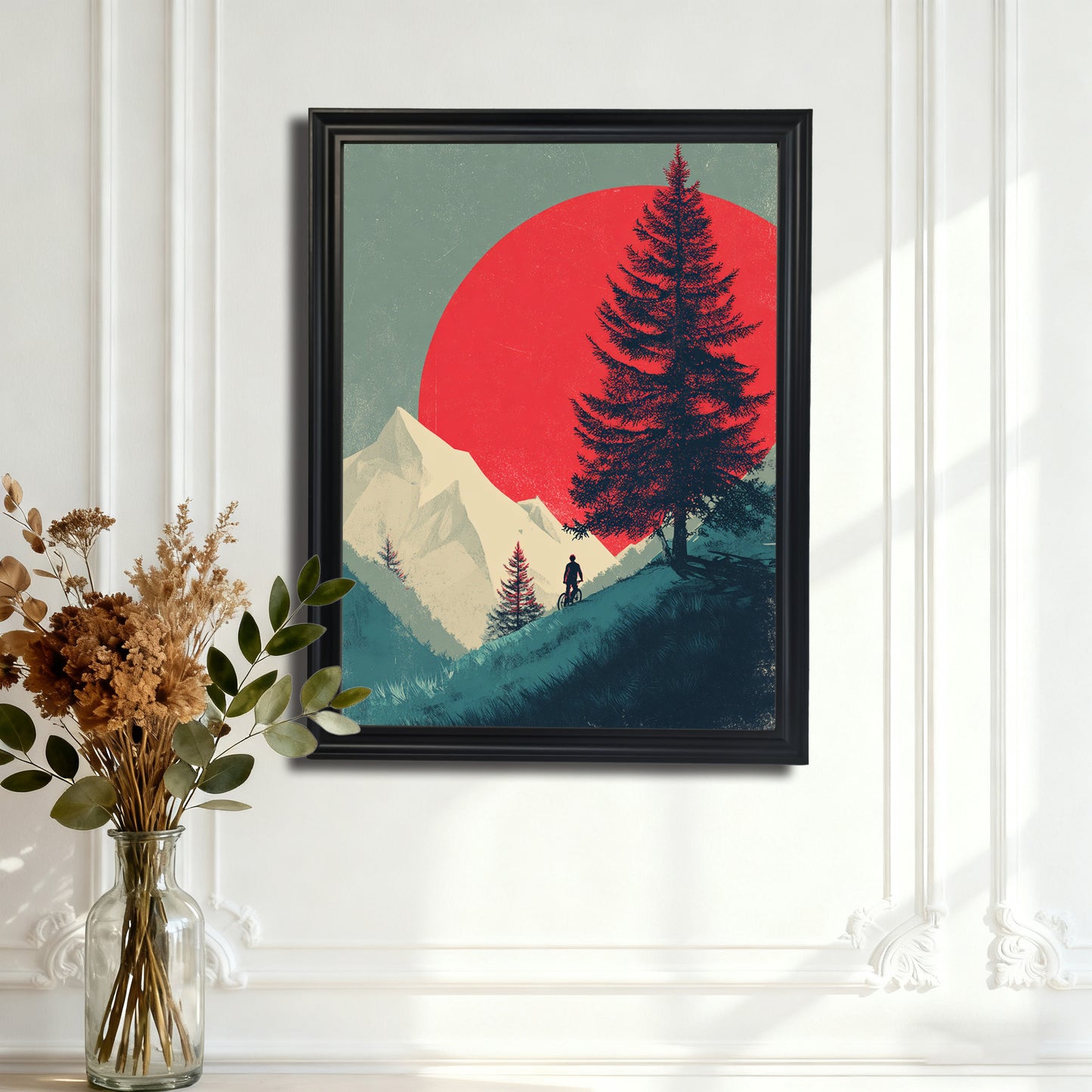 Black-framed art print of a landscape with a red sun, mountain, and pine tree silhouette, hanging on an ornate white paneled wall, with a glass vase of dried flowers in the foreground.